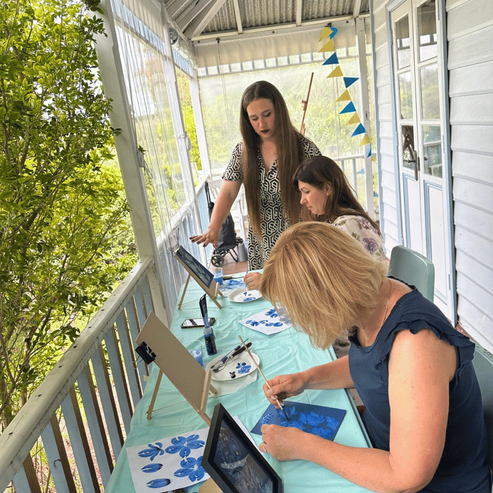 Three women sit at a table on a porch, painting blue flowers on paper. Art supplies, easels, and a blue tablecloth are visible. One woman stands, guiding the others, while the porch is decorated with blue and yellow bunting.