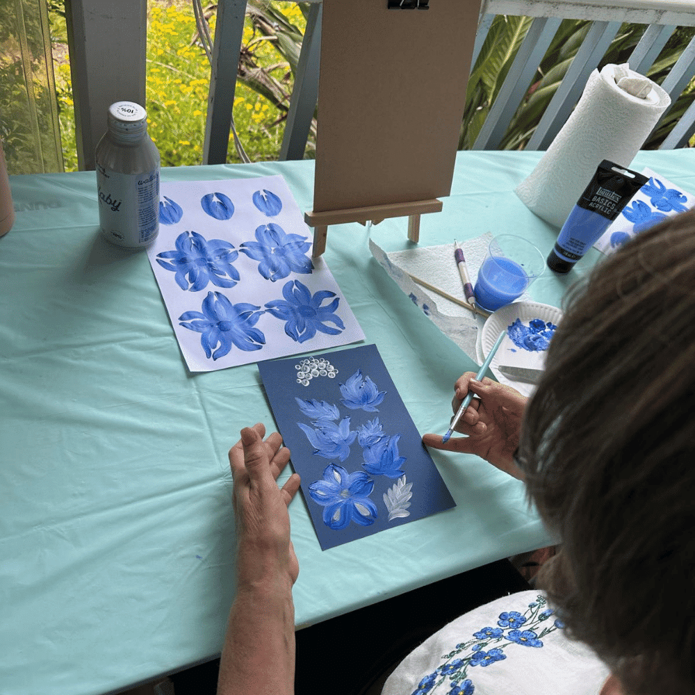 A person sits at a table painting blue and white flowers on dark paper, referencing a practice sheet. Art supplies, a small easel, bottles, and a paper towel roll are on the light blue tablecloth. Greenery is visible outside.