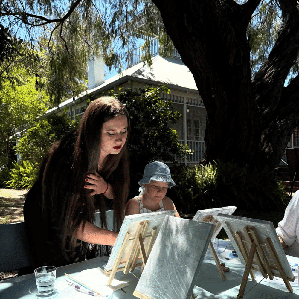 A woman stands beside a child who is painting on a small canvas at an outdoor table under a large tree, with sunlight filtering through the leaves and a house visible in the background.