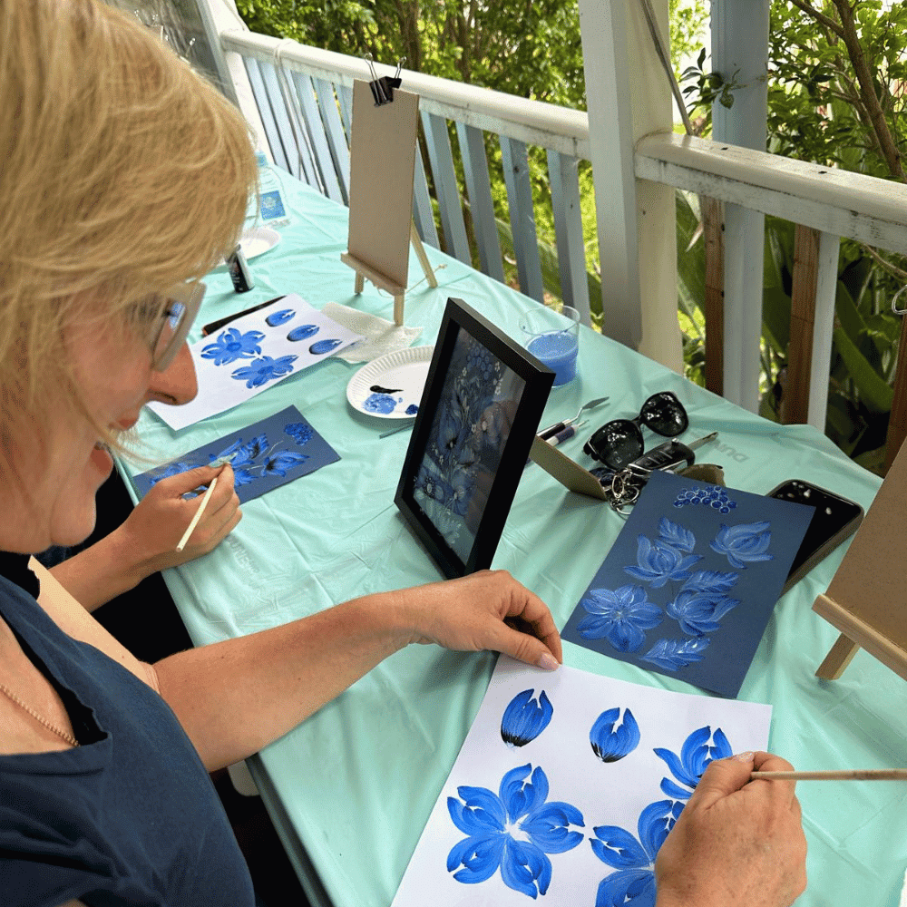 A woman smiles while painting blue flowers on white paper at an outdoor table. Finished and in-progress paintings, brushes, and an iPad displaying a reference image are also on the table.